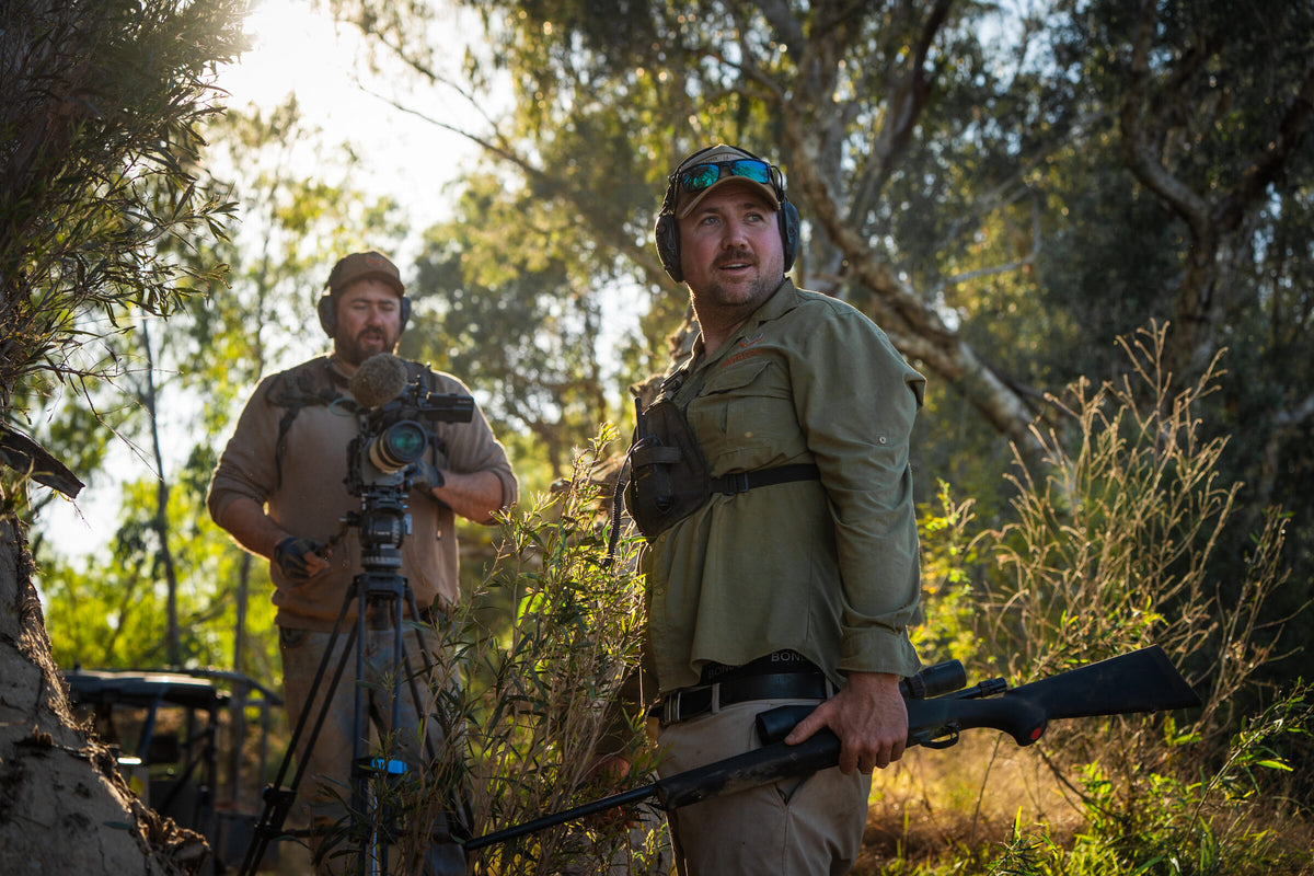 A shooter and a camera operator in backlit paperbark scrub